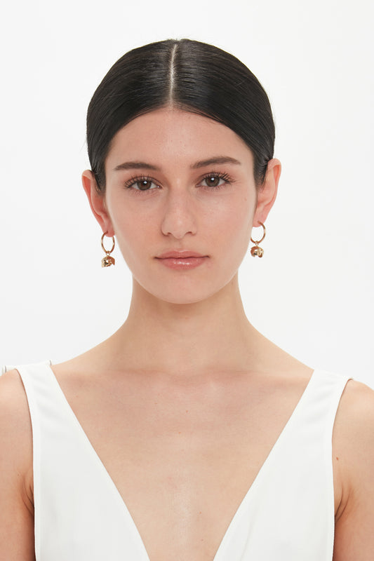 A woman with dark hair pulled back wearing Victoria Beckham's Exclusive Camellia Flower Hoop Earrings In Gold and a white V-neck top, against a plain white background.