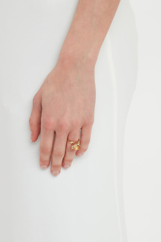 A close-up of a hand wearing an Exclusive Camellia Flower Ring In Gold by Victoria Beckham with a rectangular gemstone, handcrafted in Italy, resting against a white fabric background.