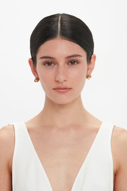 A woman with dark hair parted in the middle, wearing a white sleeveless top, poses against a plain white background. She accessorizes with Exclusive Camellia Flower Stud Earrings In Gold by Victoria Beckham, made from gold-plated brass, hand-crafted in Italy.