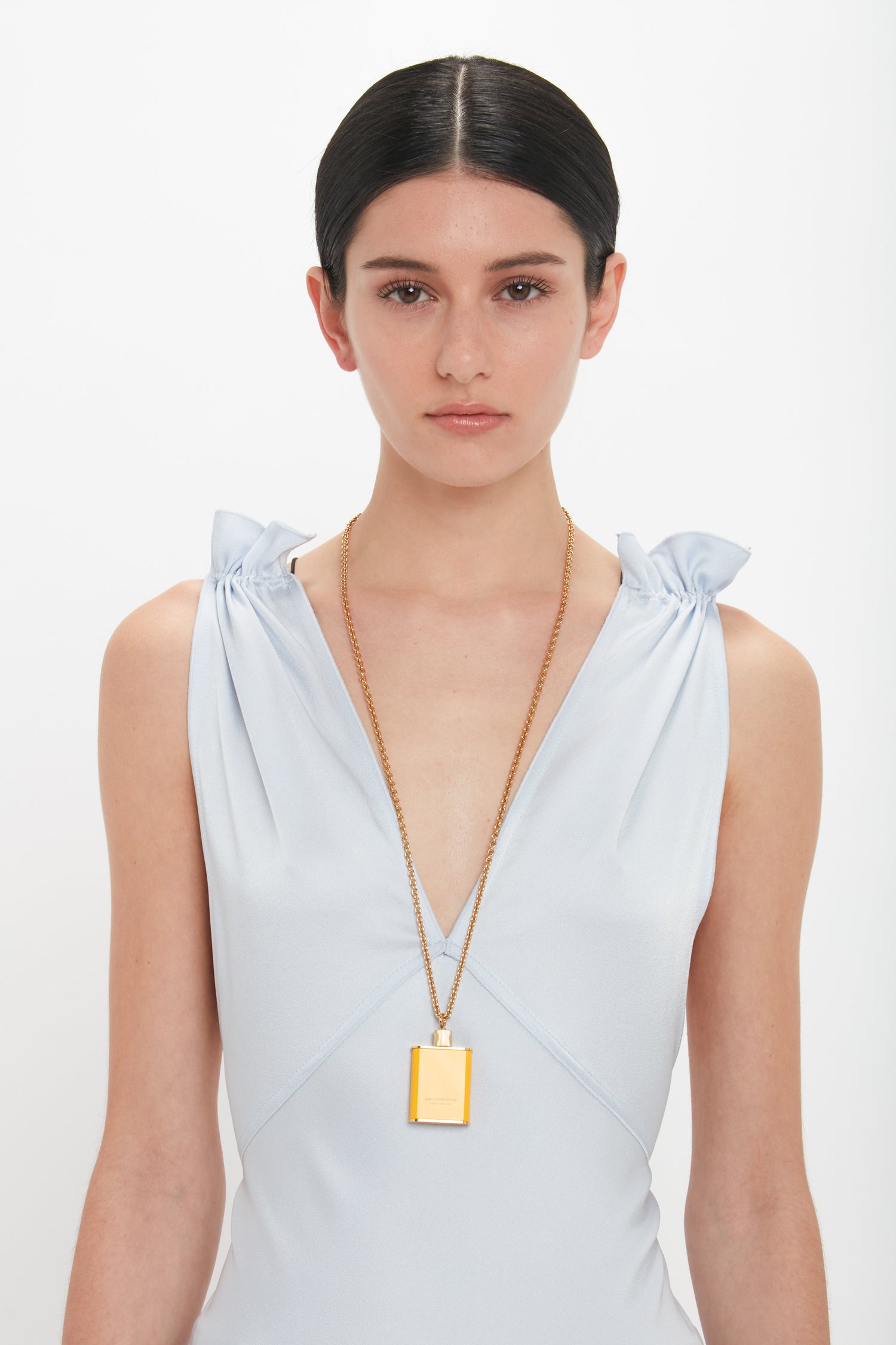 Woman in a light blue sleeveless dress, with minimal makeup and straight hair, wearing a long gold brushed brass Perfume Bottle Necklace In San Ysidro Drive by Victoria Beckham. She is standing against a white background.