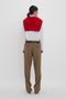 Person standing against a white background, facing away. Wearing a white shirt, Victoria Beckham Asymmetric Chino Trouser In Tobacco with a narrow leg silhouette, and a red sweater draped over the shoulders, tied in the front.