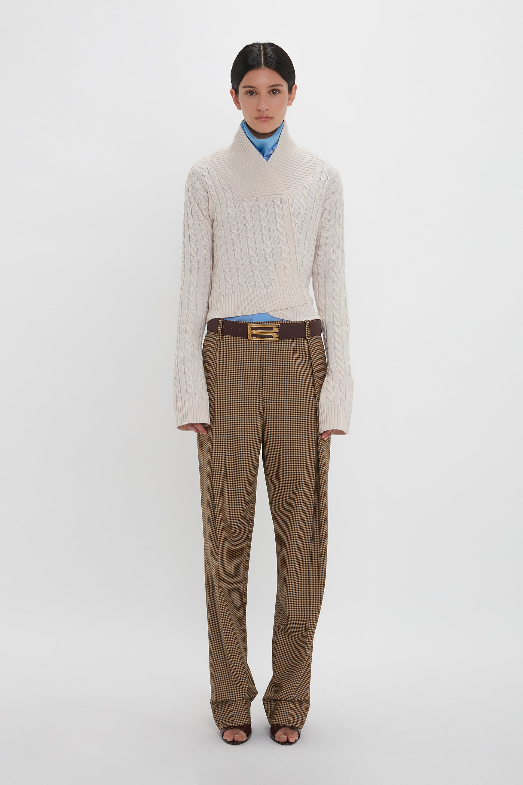 Person standing against a plain background, wearing a Wrap Detail Jumper In Bone by Victoria Beckham, brown plaid trousers, blue shirt underneath, and brown belt. They are looking straight ahead.