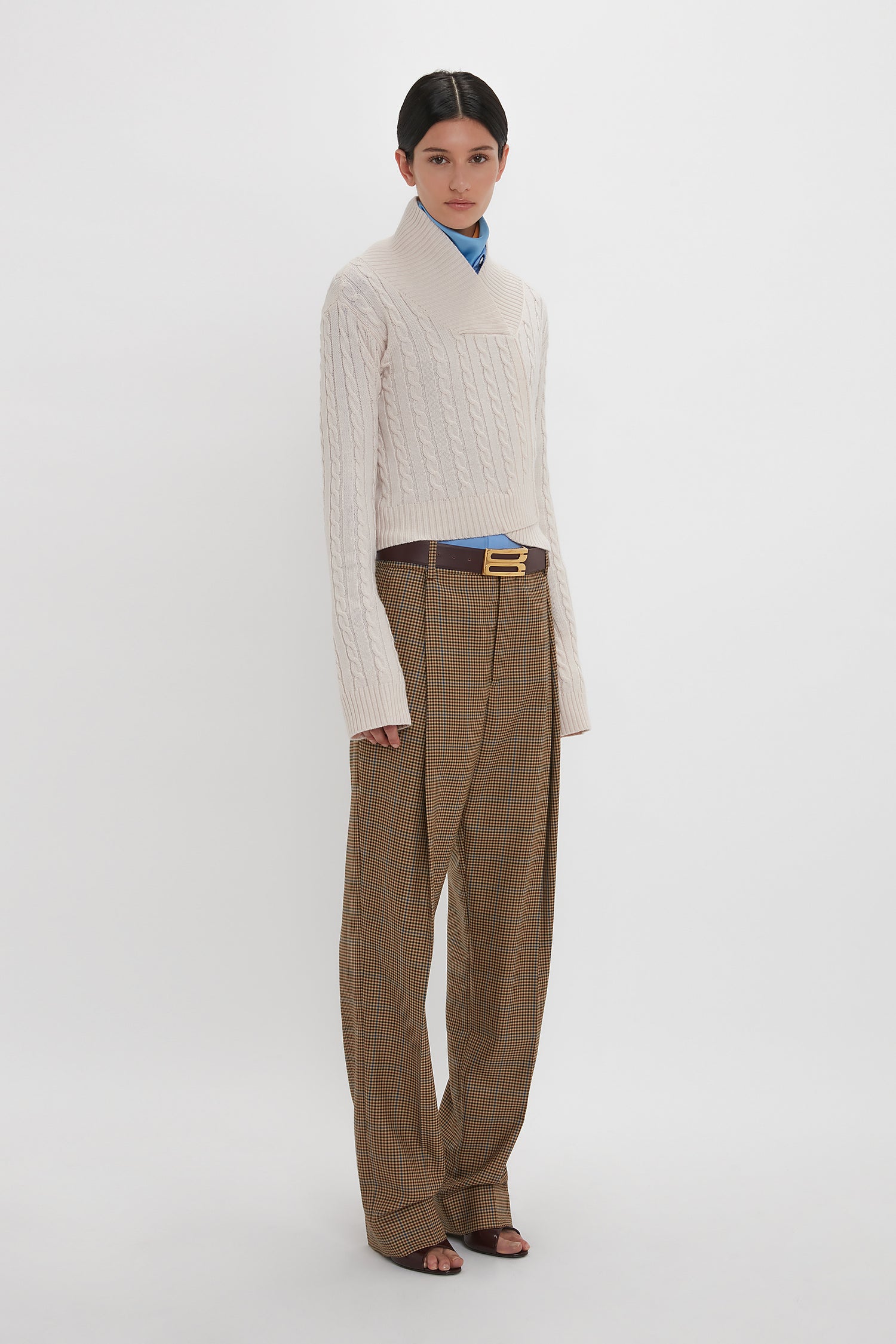 A person stands against a plain background wearing a Wrap Detail Jumper In Bone by Victoria Beckham, paired with a blue collared shirt, brown plaid wide-leg trousers, and black sandals.