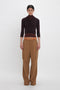 A person with a neutral expression is wearing a dark long-sleeve top and Relaxed Cargo Trouser In Tobacco by Victoria Beckham with a tan belt, standing against a plain white background. The outfit showcases a relaxed silhouette, perfect for everyday wear.