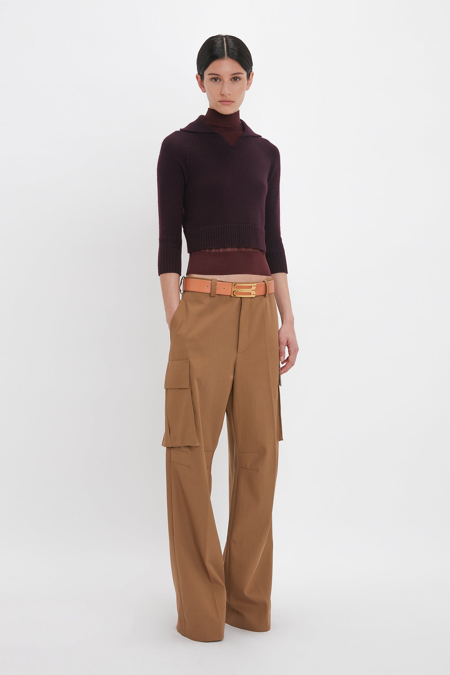 Person standing against a white background wearing a dark maroon top, Relaxed Cargo Trouser In Tobacco by Victoria Beckham, and a beige belt with a gold buckle, showcasing a military-inspired style with a relaxed silhouette.