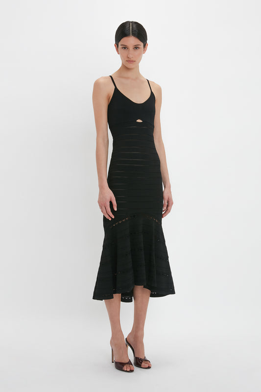 Person wearing a sleeveless black Cut-Out Detail Cami Dress In Black by Victoria Beckham with spaghetti straps and a small cut-out detail at the front, standing against a white background. They are also wearing black high-heeled shoes.