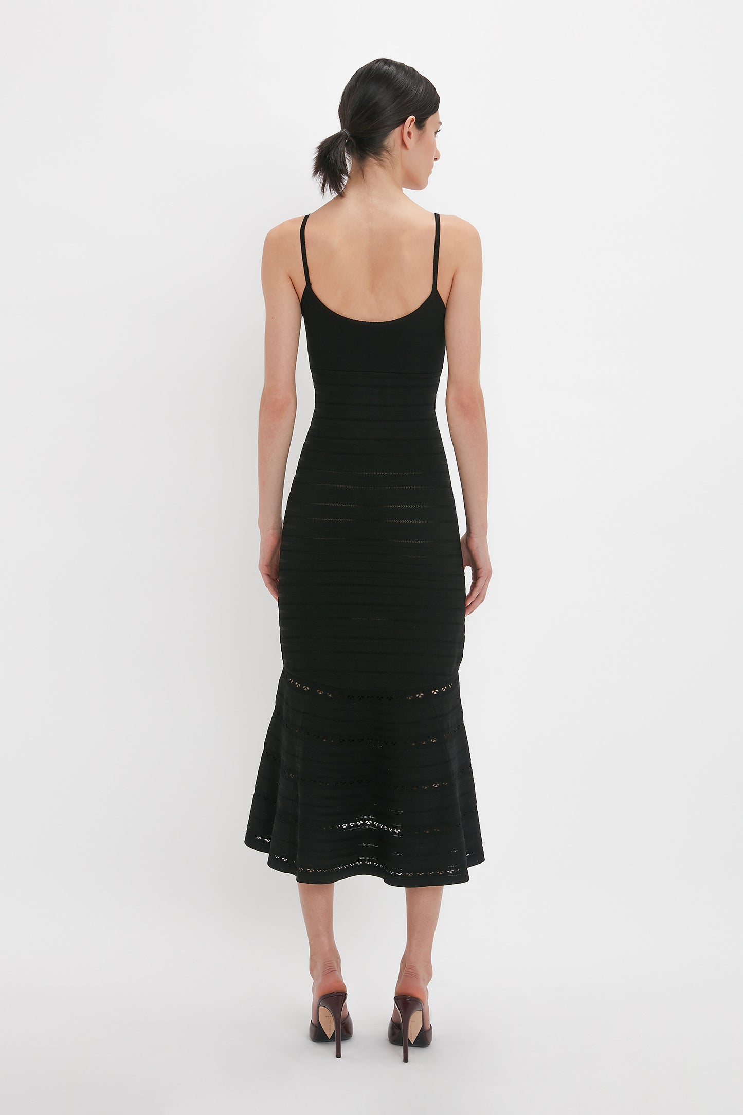 A person with dark hair tied back is standing with their back to the camera, wearing a Victoria Beckham Cut-Out Detail Cami Dress In Black and black high-heeled shoes.