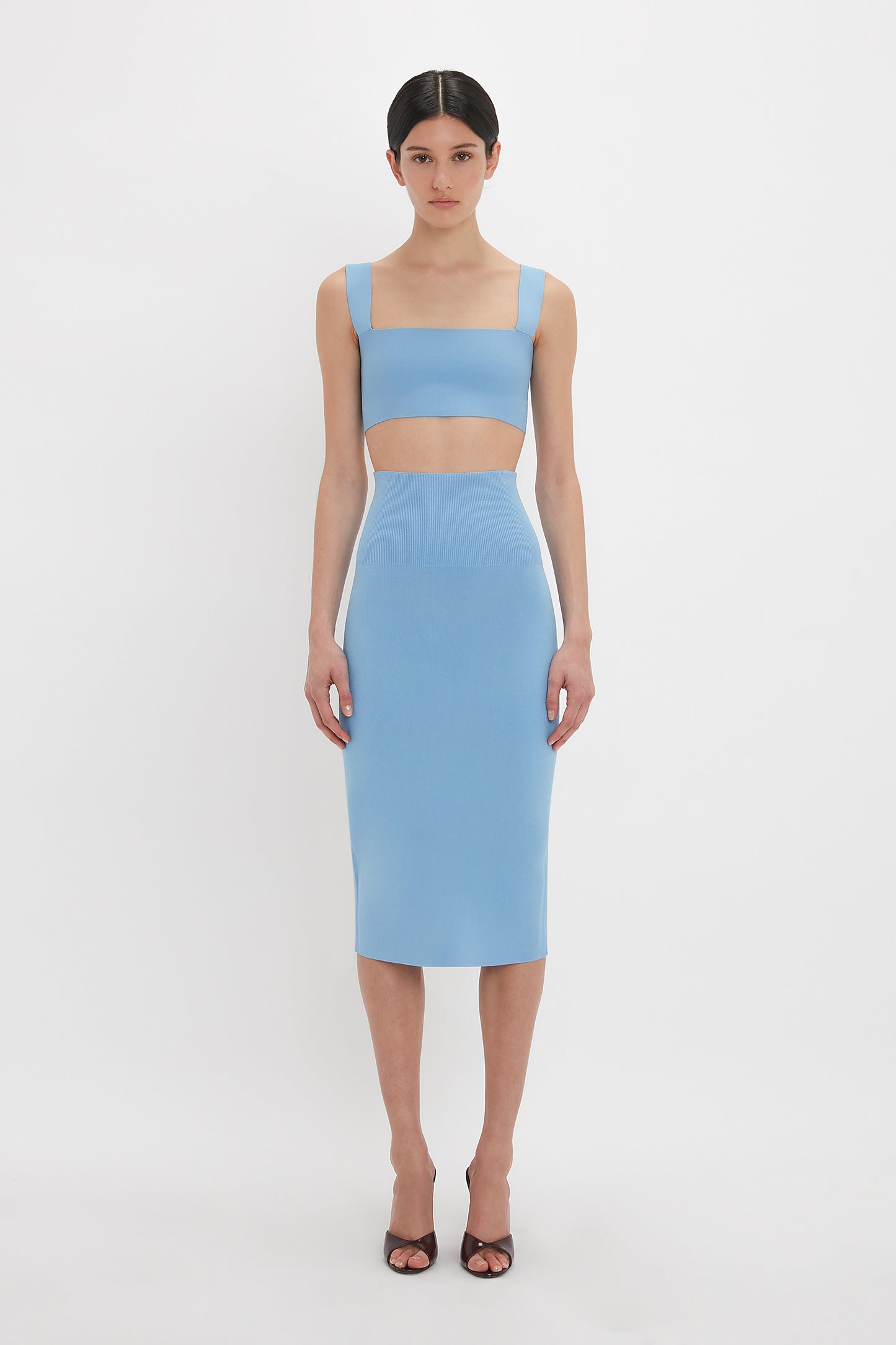 A person stands against a plain white background wearing the Victoria Beckham Strap Bandeau Top In Marina with a compact knitwear light blue two-piece outfit with wide straps and a knee-length skirt, along with open-toe heels.