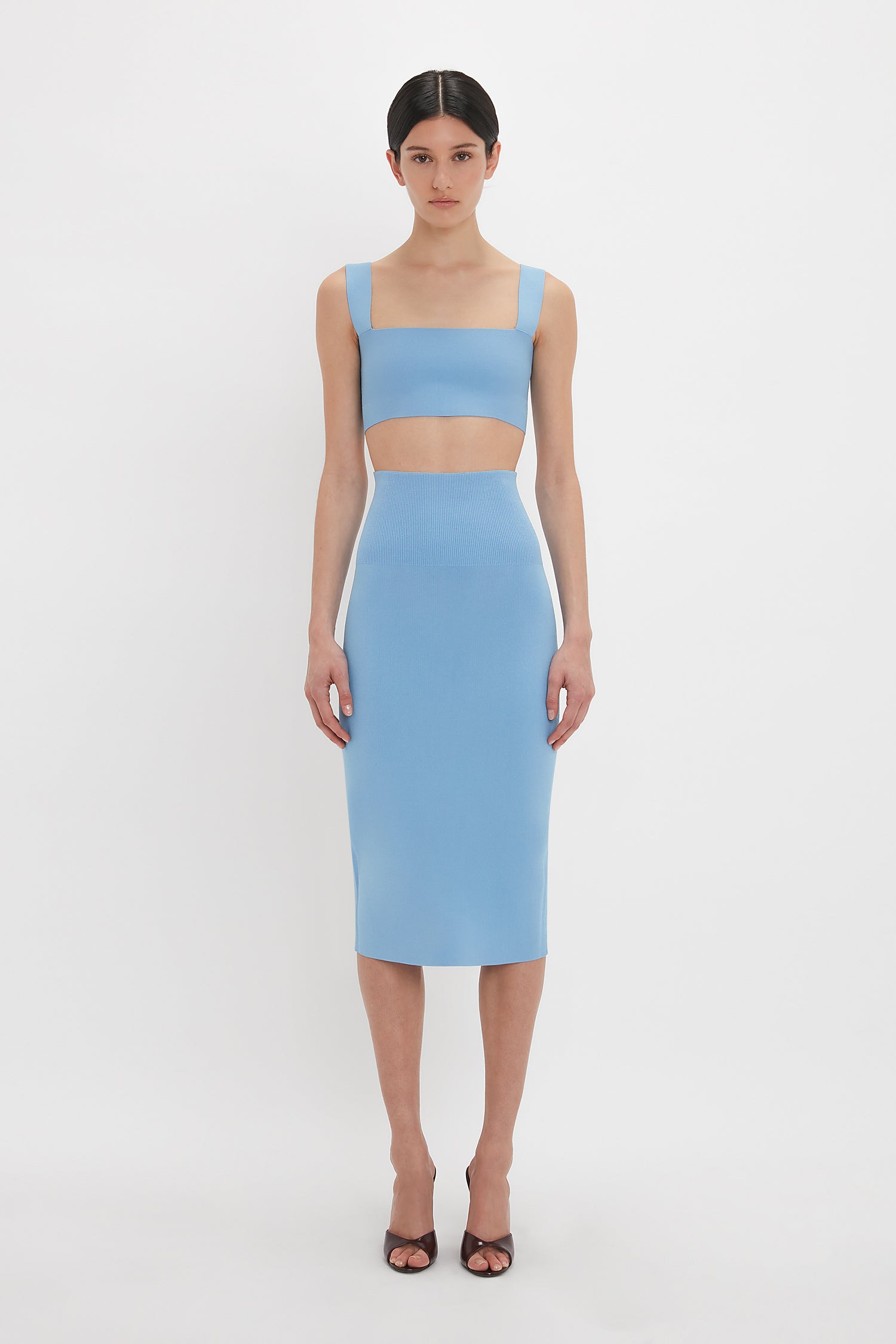 A person stands against a plain white background wearing the Victoria Beckham Strap Bandeau Top In Marina with a compact knitwear light blue two-piece outfit with wide straps and a knee-length skirt, along with open-toe heels.