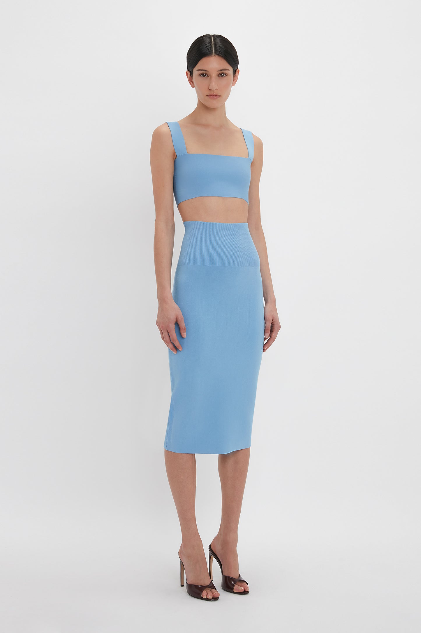 A woman stands against a white background, wearing an ultra form-fitting, light blue two-piece outfit consisting of the Victoria Beckham Strap Bandeau Top In Marina and a high-waisted knee-length skirt. She is also wearing black high-heeled sandals.