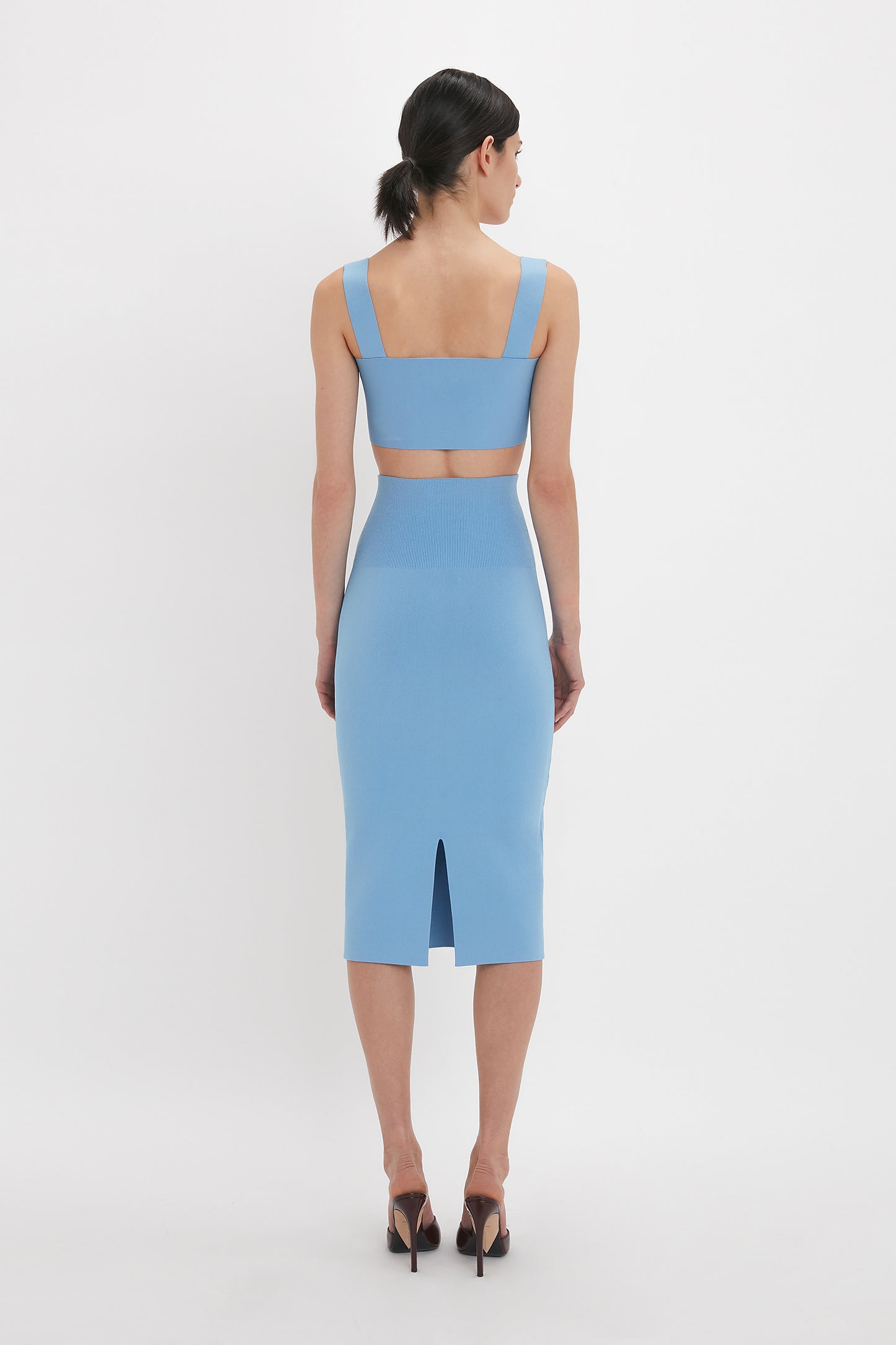 A person with dark hair pulled back stands facing away, wearing a blue sleeveless dress, compact knitwear, black high heels, and a Strap Bandeau Top In Marina by Victoria Beckham.