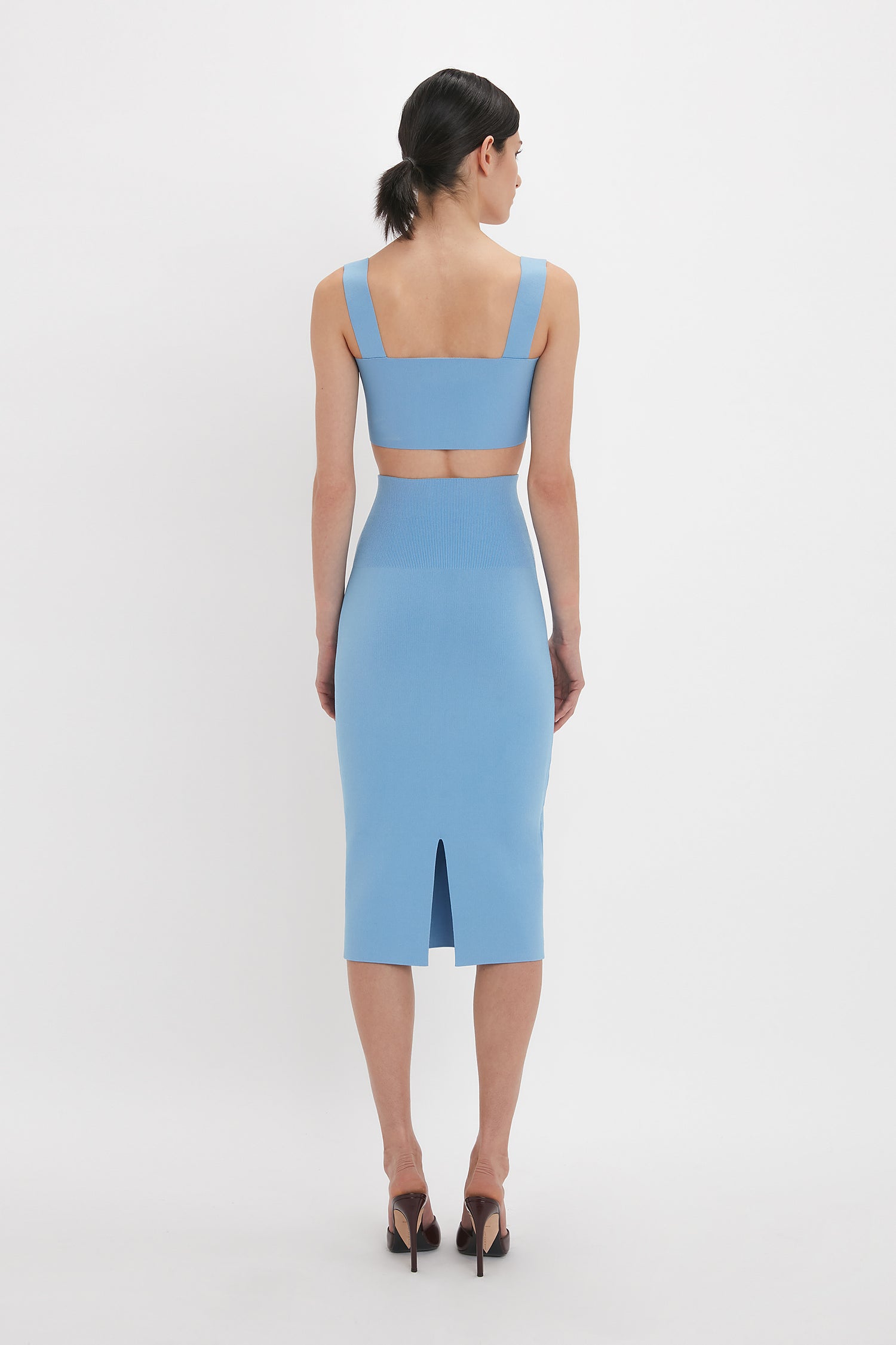 A woman with black hair tied back is wearing a contemporary polished blue sleeveless dress with a cut-out back and a slit at the bottom, standing against a plain white background, featuring the Fitted Midi Skirt In Marina by Victoria Beckham.