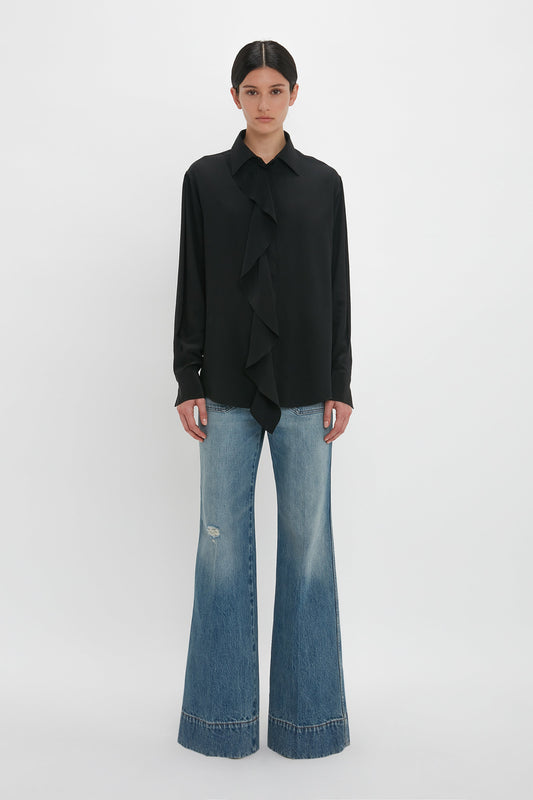 Person wearing a classic Victoria Beckham Asymmetric Ruffle Blouse In Black with light-washed flare jeans, standing against a plain white background.