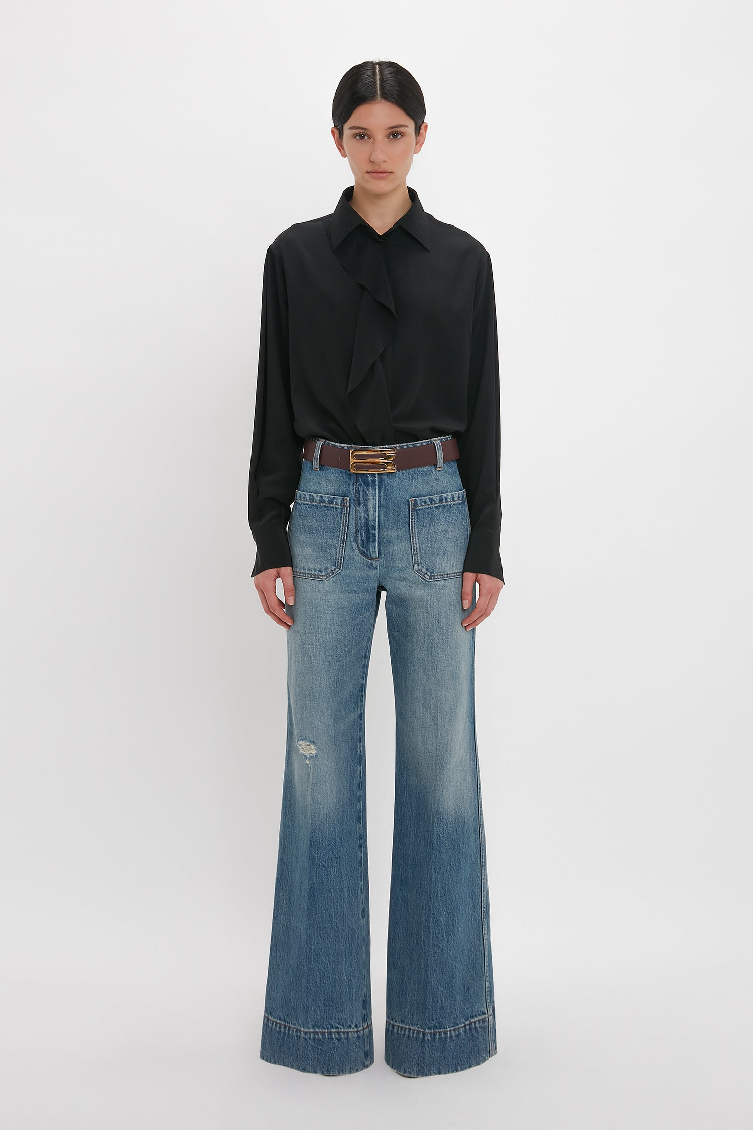 A person stands against a plain white background, wearing a black blouse with a ruffled neckline, Victoria Beckham Alina High Waisted Jean In Worn Blue Wash with seventies-inspired patch pockets, and a brown belt.