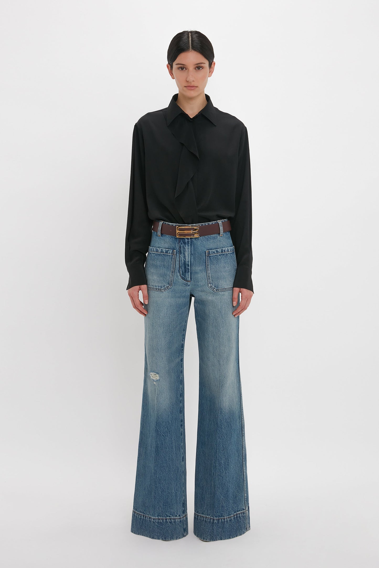 A person stands against a plain background, wearing a Victoria Beckham Asymmetric Ruffle Blouse In Black, blue wide-leg jeans with large front pockets, and a belt.