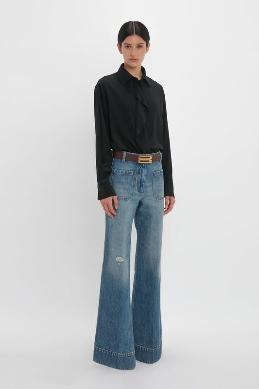 A person stands against a plain white background, wearing the Victoria Beckham Asymmetric Ruffle Blouse In Black paired with high-waisted wide-leg jeans with a belt.