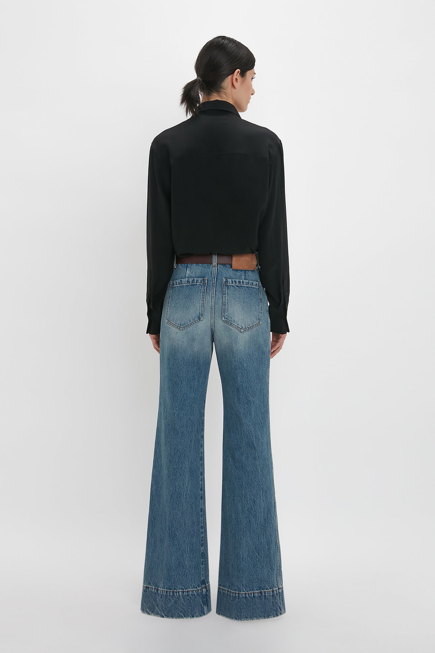 A person with dark hair tied back stands facing away, wearing a black long-sleeved shirt and Victoria Beckham's Alina High Waisted Jean In Worn Blue Wash featuring seventies-inspired patch pockets. The background is plain white.