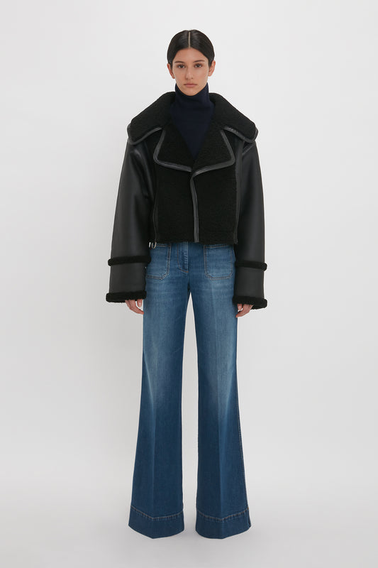 A person stands in a studio wearing a Shearling Jacket In Black by Victoria Beckham over a black turtleneck and wide-leg blue jeans.