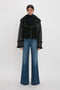 A person stands in a studio wearing a Shearling Jacket In Black by Victoria Beckham over a black turtleneck and wide-leg blue jeans.