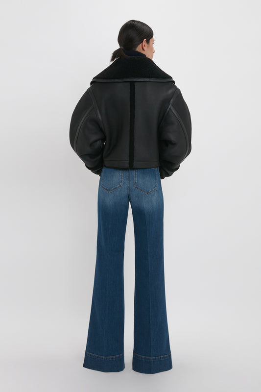 A person with dark hair stands facing away, wearing a Shearling Jacket In Black by Victoria Beckham and wide-legged blue jeans against a plain white background.