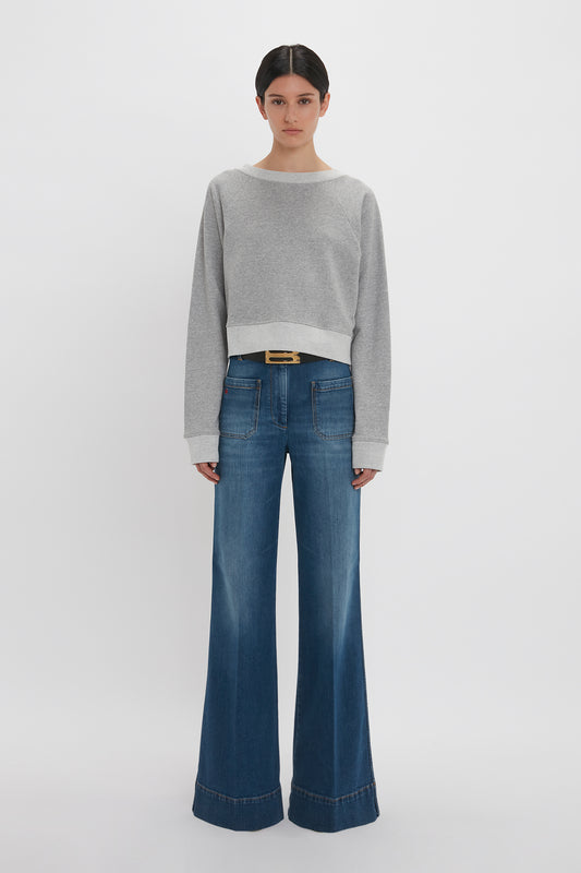 A woman in a Victoria Beckham Cropped Sweatshirt In Grey Marl and flared blue jeans stands against a plain white backdrop.