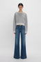 A woman in a Victoria Beckham Cropped Sweatshirt In Grey Marl and flared blue jeans stands against a plain white backdrop.