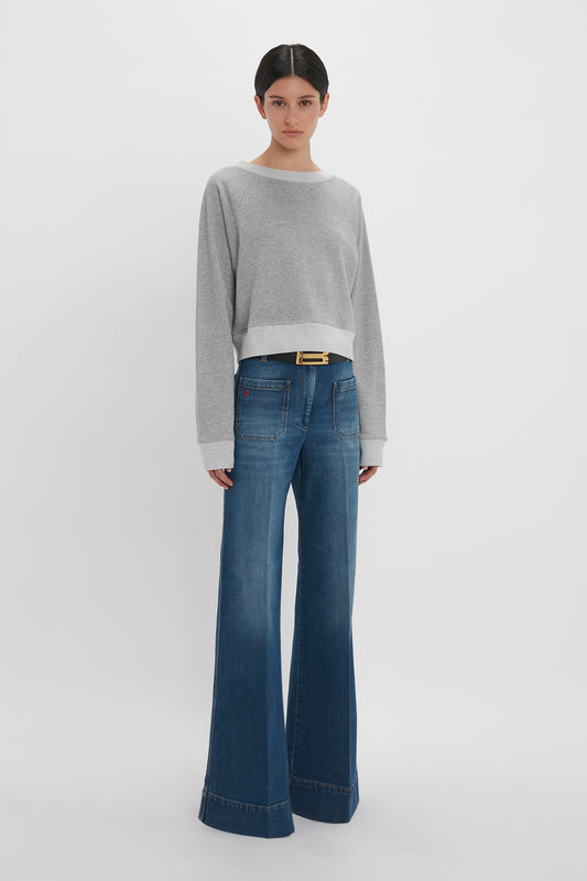 A woman standing in a white studio, wearing a Victoria Beckham grey marl cropped sweatshirt and blue flared jeans with a slim black belt.