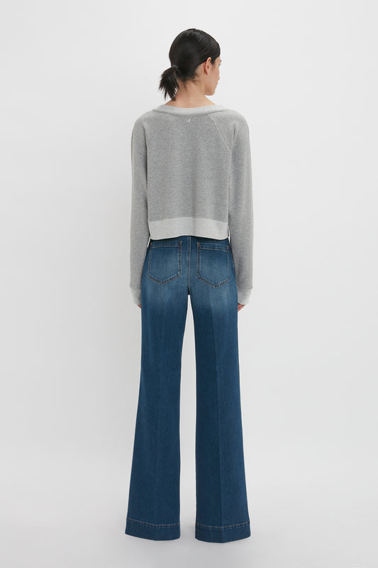Woman standing facing away, wearing a Victoria Beckham grey marl cropped sweatshirt and blue flared jeans, against a white background.