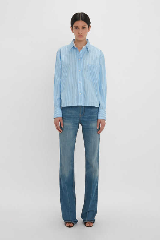 A person with a neutral expression is wearing a light blue, **Cropped Long Sleeve Shirt In Marina-White by Victoria Beckham** and blue jeans, standing against a plain white background.