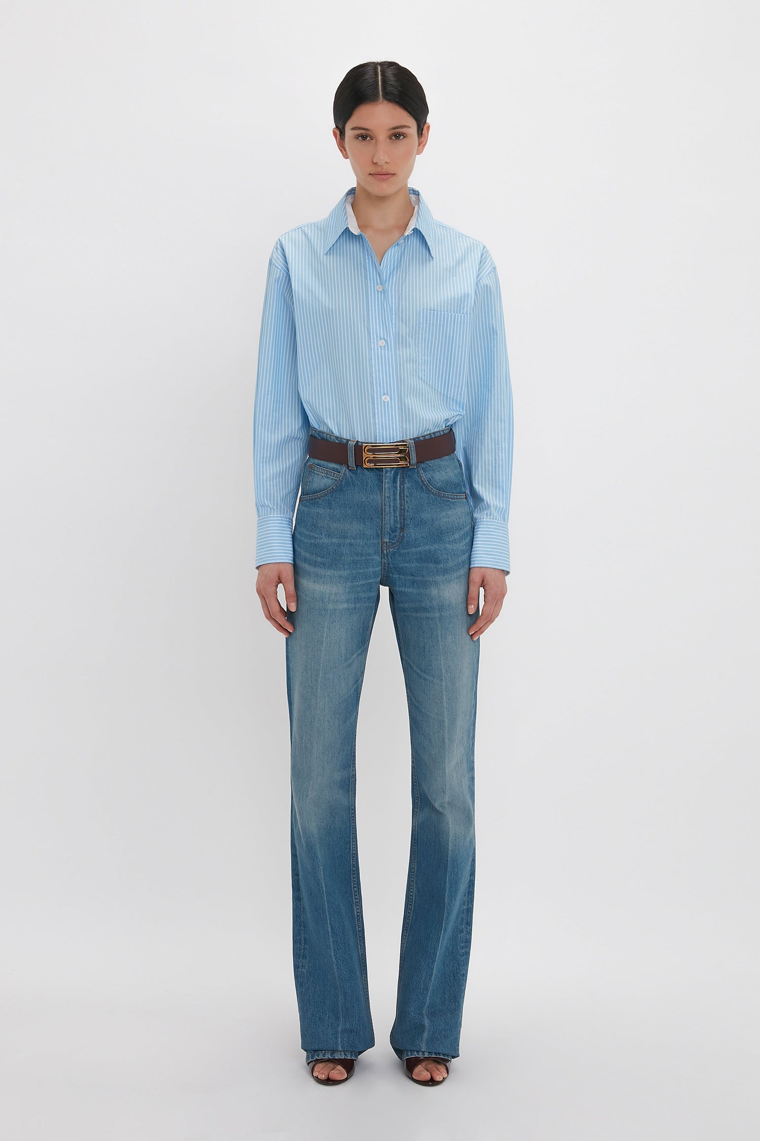 A person stands facing forward, wearing a Victoria Beckham Cropped Long Sleeve Shirt In Marina-White, blue jeans, and a brown belt. They have their hair pulled back and are posing against a white background.