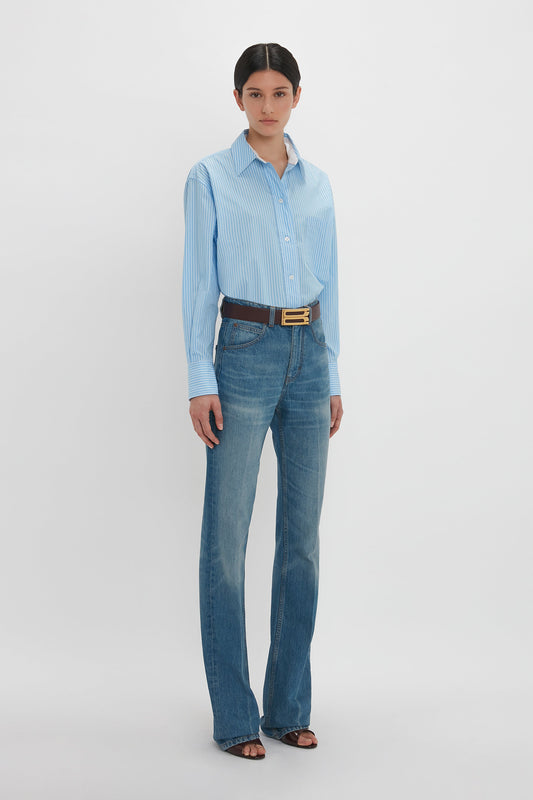 A person stands against a plain background wearing a light blue striped Cropped Long Sleeve Shirt In Marina-White by Victoria Beckham, blue jeans, a brown belt, and black shoes.