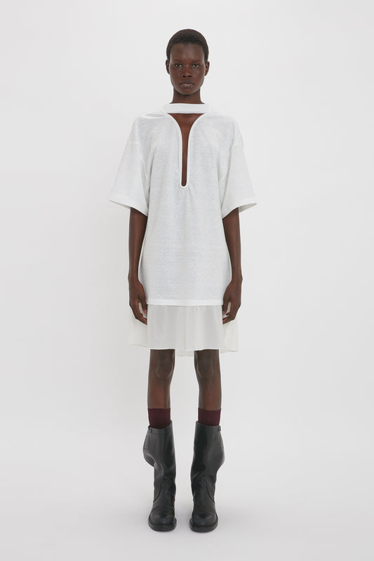 A person stands against a plain white background, wearing a Frame Cut-Out T-Shirt Dress In White by Victoria Beckham and black boots.
