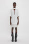 A person stands against a plain white background, wearing a Frame Cut-Out T-Shirt Dress In White by Victoria Beckham and black boots.