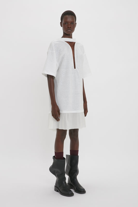A person stands against a plain background wearing a loose, short-sleeved Frame Cut-Out T-Shirt Dress In White by Victoria Beckham with a navel-grazing keyhole neckline and black knee-high boots.