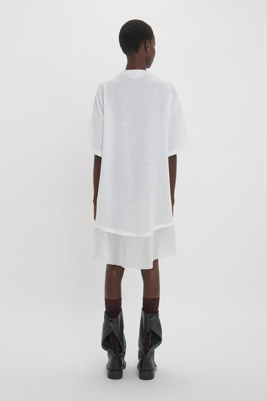 A person is standing with their back to the camera, wearing a Victoria Beckham Frame Cut-Out T-Shirt Dress In White, a white skirt, and black boots.