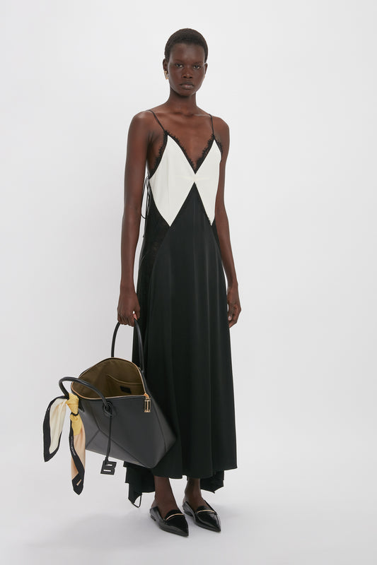A person stands against a white background wearing a black and white V-Neck Lace Insert Cami Dress In Ivory by Victoria Beckham with retro 1990s slip dress vibes, holding a large black bag with a patterned scarf attached to it.