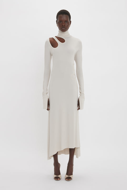 Person standing against a white background wearing a Victoria Beckham Long Sleeve Cut-Out Jersey Midi Dress In Bone with a high neck, asymmetric hemline, and asymmetrical cutouts.
