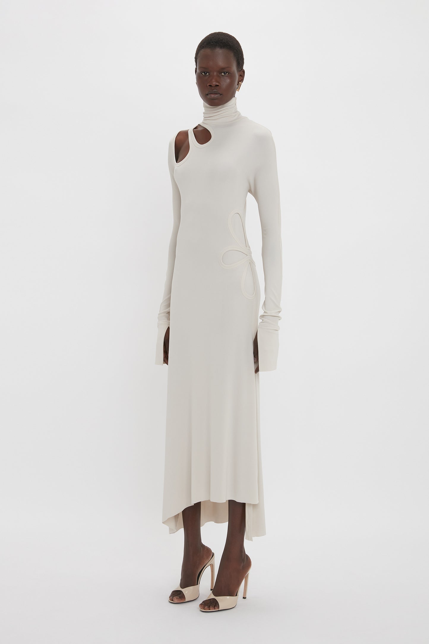 Person wearing a Victoria Beckham Long Sleeve Cut-Out Jersey Midi Dress In Bone with a high neckline and an asymmetric hemline, paired with high-heeled shoes, standing against a plain white background.