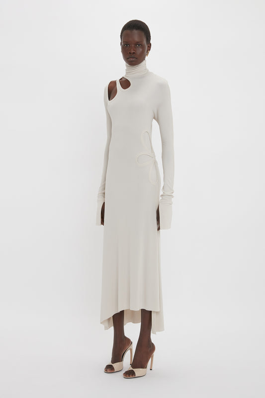Person wearing a Victoria Beckham Long Sleeve Cut-Out Jersey Midi Dress In Bone with a high neckline and an asymmetric hemline, paired with high-heeled shoes, standing against a plain white background.