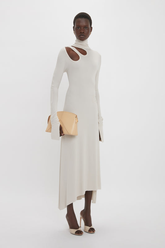 A person wearing a Victoria Beckham Long Sleeve Cut-Out Jersey Midi Dress In Bone with an asymmetric hemline, holding a beige clutch purse, and standing against a plain white background.