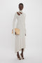 A person wearing a Victoria Beckham Long Sleeve Cut-Out Jersey Midi Dress In Bone with an asymmetric hemline, holding a beige clutch purse, and standing against a plain white background.