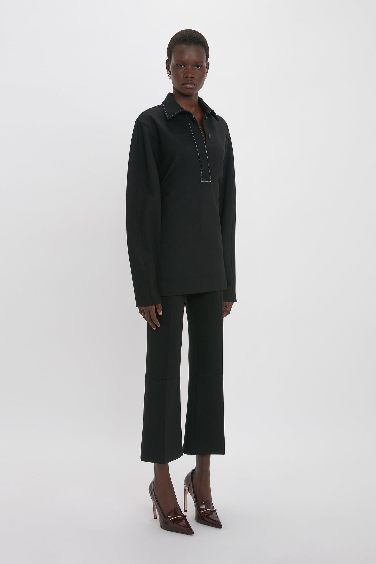 A person stands facing forward, exuding a distinctive personality in the Waistcoat Detail Ponti Top In Black by Victoria Beckham, black pants, and dark high-heeled shoes against a white background.