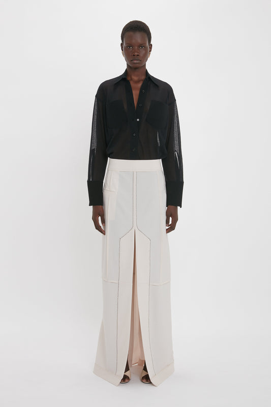 A person stands against a white background, wearing the Pocket Detail Shirt In Black by Victoria Beckham with a collared neckline and a high-waisted, long white skirt with a front slit. The person is looking directly at the camera.