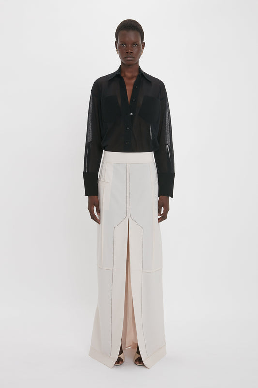 A person stands against a plain background wearing a sheer black shirt with exposed seams and the Victoria Beckham Deconstructed Floor-Length Skirt In Bone, embodying relaxed refinement.