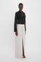 Person standing against a white background, wearing the Pocket Detail Shirt In Black by Victoria Beckham and a long white skirt with a slit in the front.