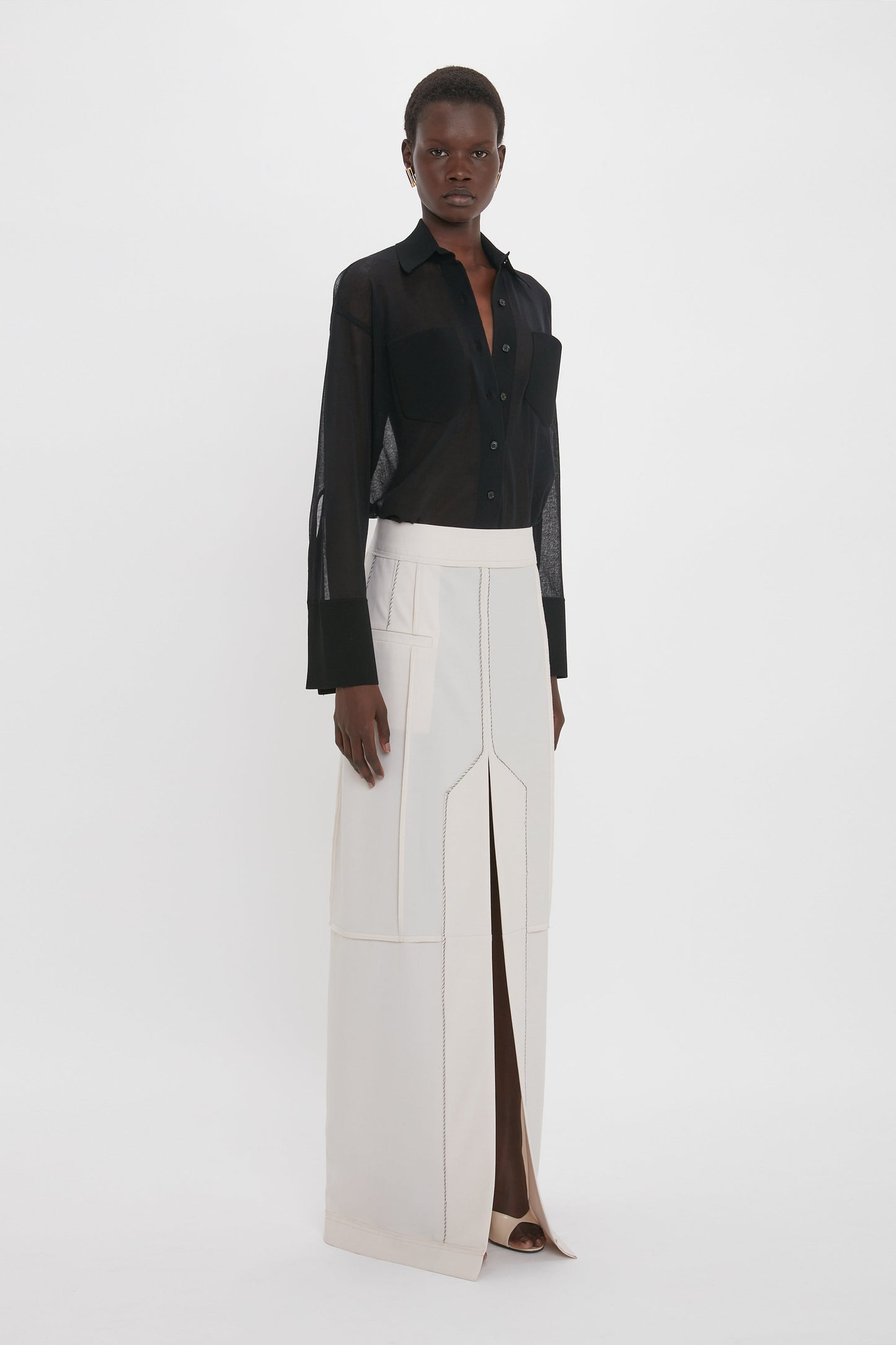 Person standing against a plain background wearing a sheer black button-down shirt and a long, high-waisted Deconstructed Floor-Length Skirt In Bone by Victoria Beckham with exposed seams and a front slit, exuding relaxed refinement.