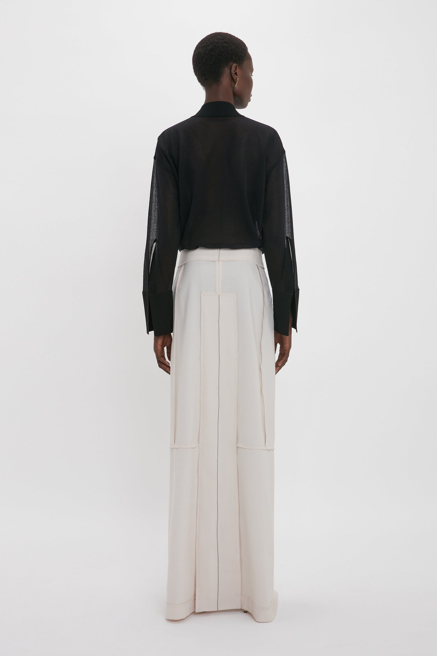 A person is standing with their back to the camera, wearing a black long-sleeve top and the Victoria Beckham Deconstructed Floor-Length Skirt In Bone that conveys a sense of relaxed refinement in front of a plain white background.