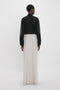 A person is standing with their back to the camera, wearing a black long-sleeve top and the Victoria Beckham Deconstructed Floor-Length Skirt In Bone that conveys a sense of relaxed refinement in front of a plain white background.