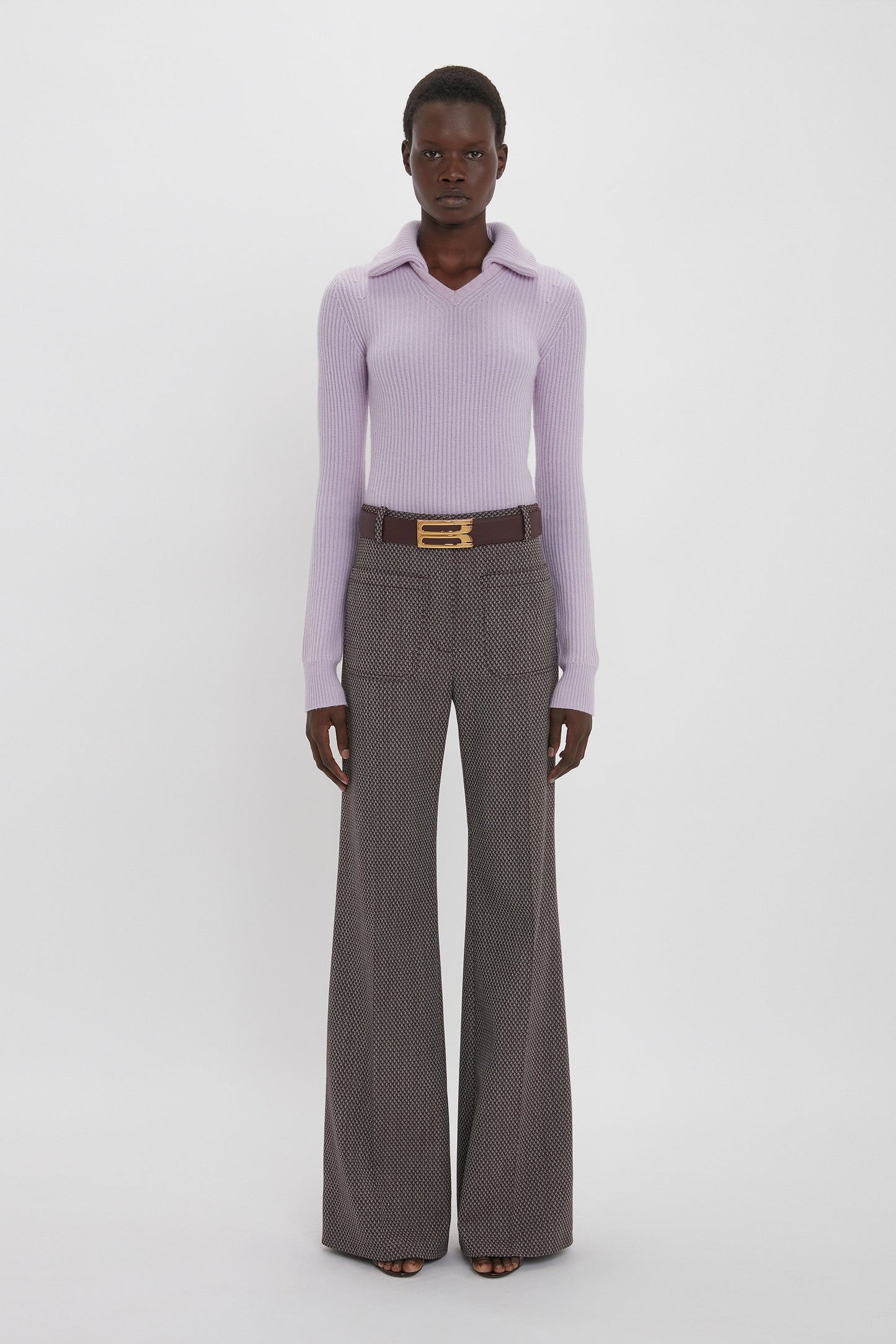 A person stands against a plain background, wearing a long-sleeved lavender top, Alina Trouser In Tobacco by Victoria Beckham, and a belt with a gold buckle.
