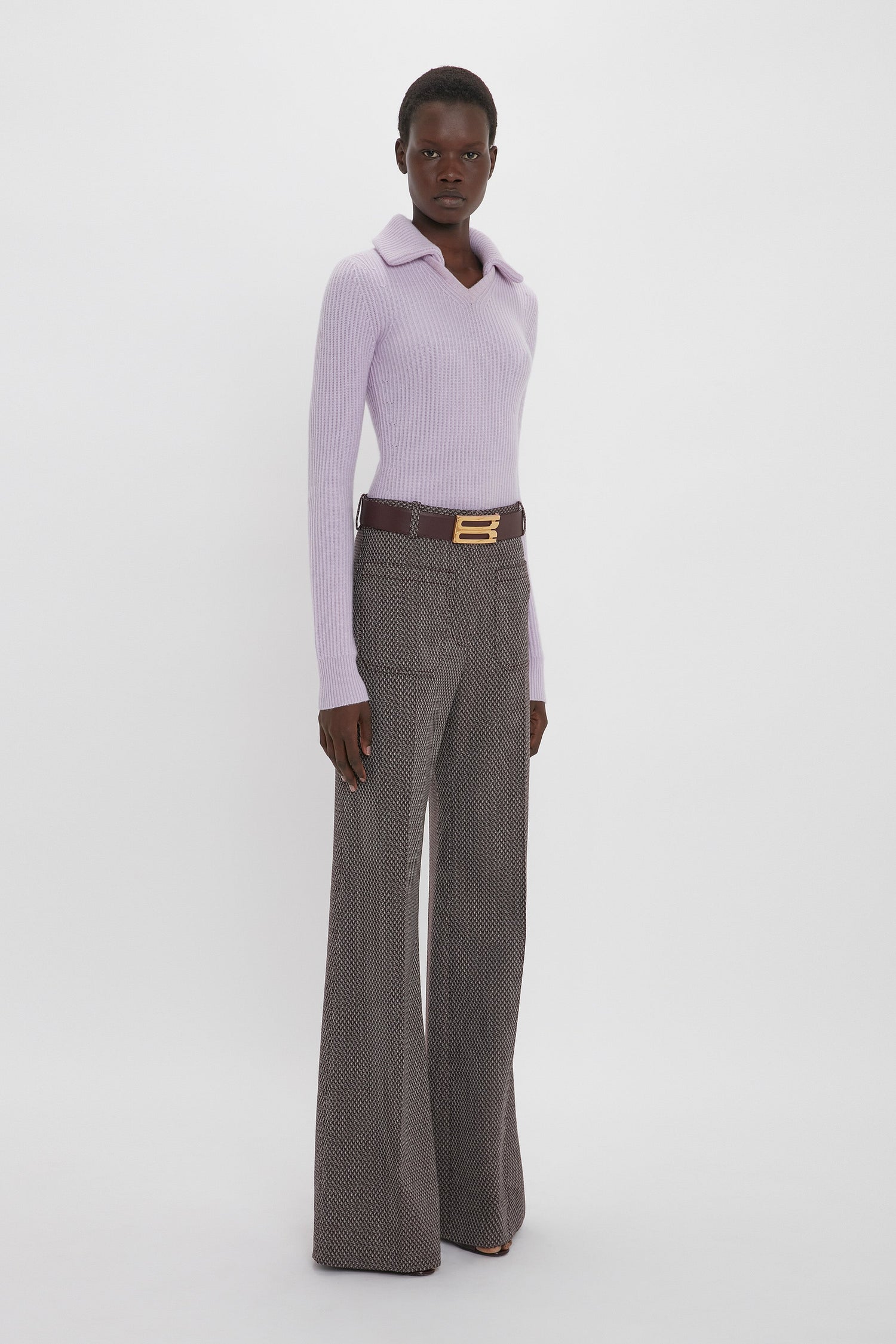 A person stands against a plain background, wearing a light purple sweater with a collar and Victoria Beckham's Alina Trouser In Tobacco featuring flared legs and a belt with a gold buckle.